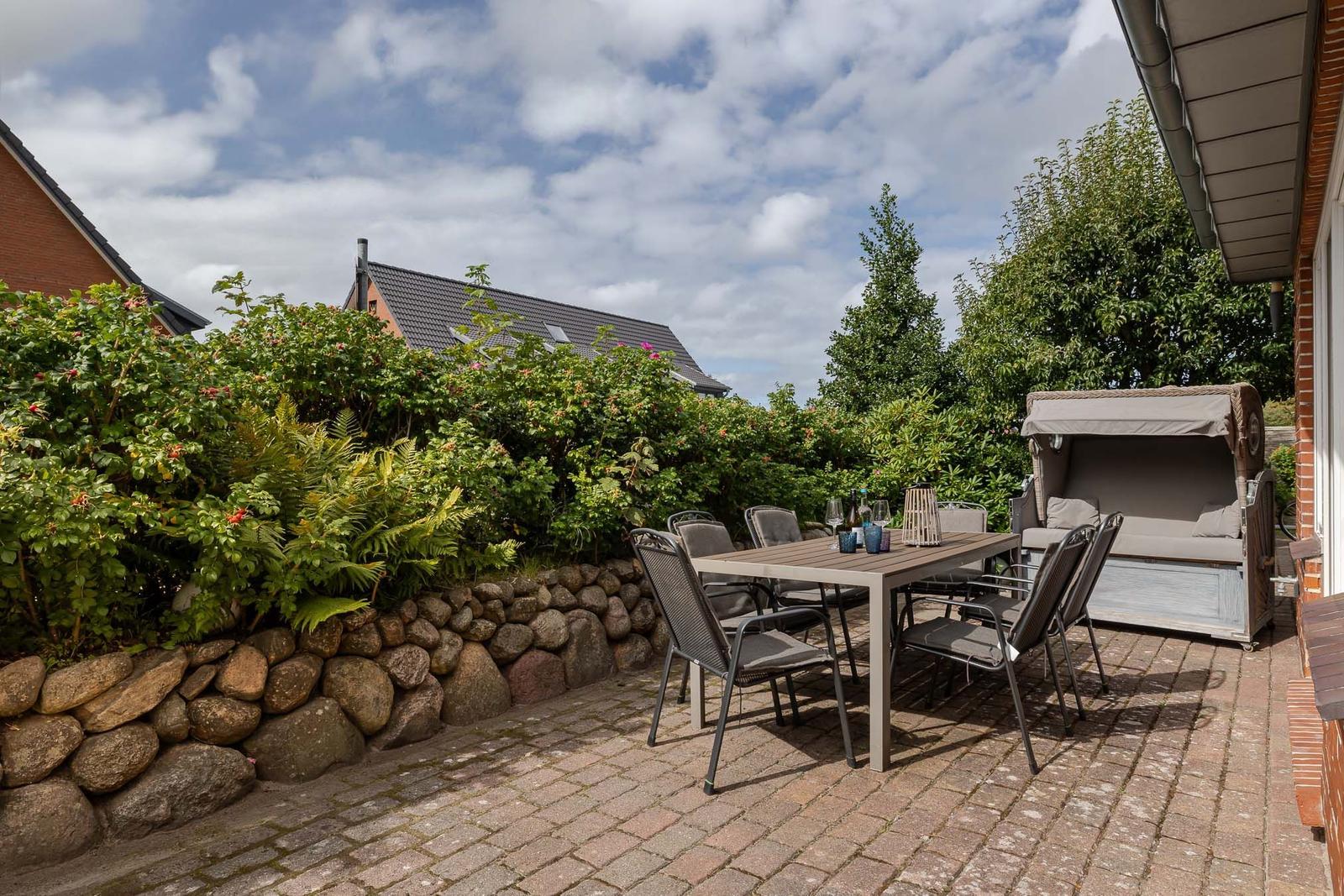 Pflasterterrasse mit Esstisch, sechs Stühlen, Strandkorb und niedriger Steinmauer.