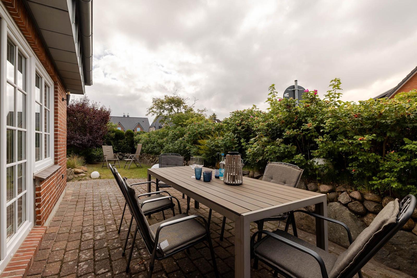 Terrasse mit Holztisch, grauen Gartenstühlen, Pflaster und grüner Hecke.