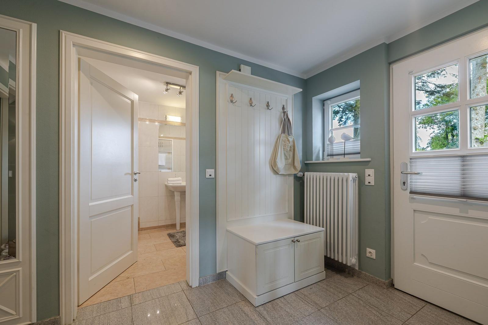 Flur mit weißer Garderobe, offener Badtür, Heizkörper und Fenster mit Plissee.