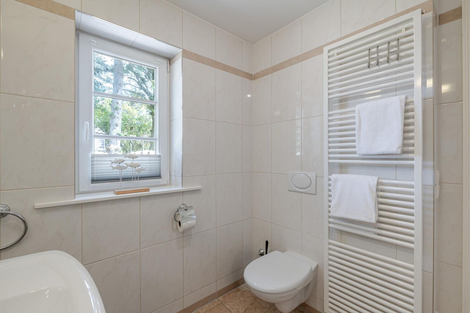 Hell gefliestes Bad mit Wand-WC, Handtuchheizkörper und Fenster mit Plissee.