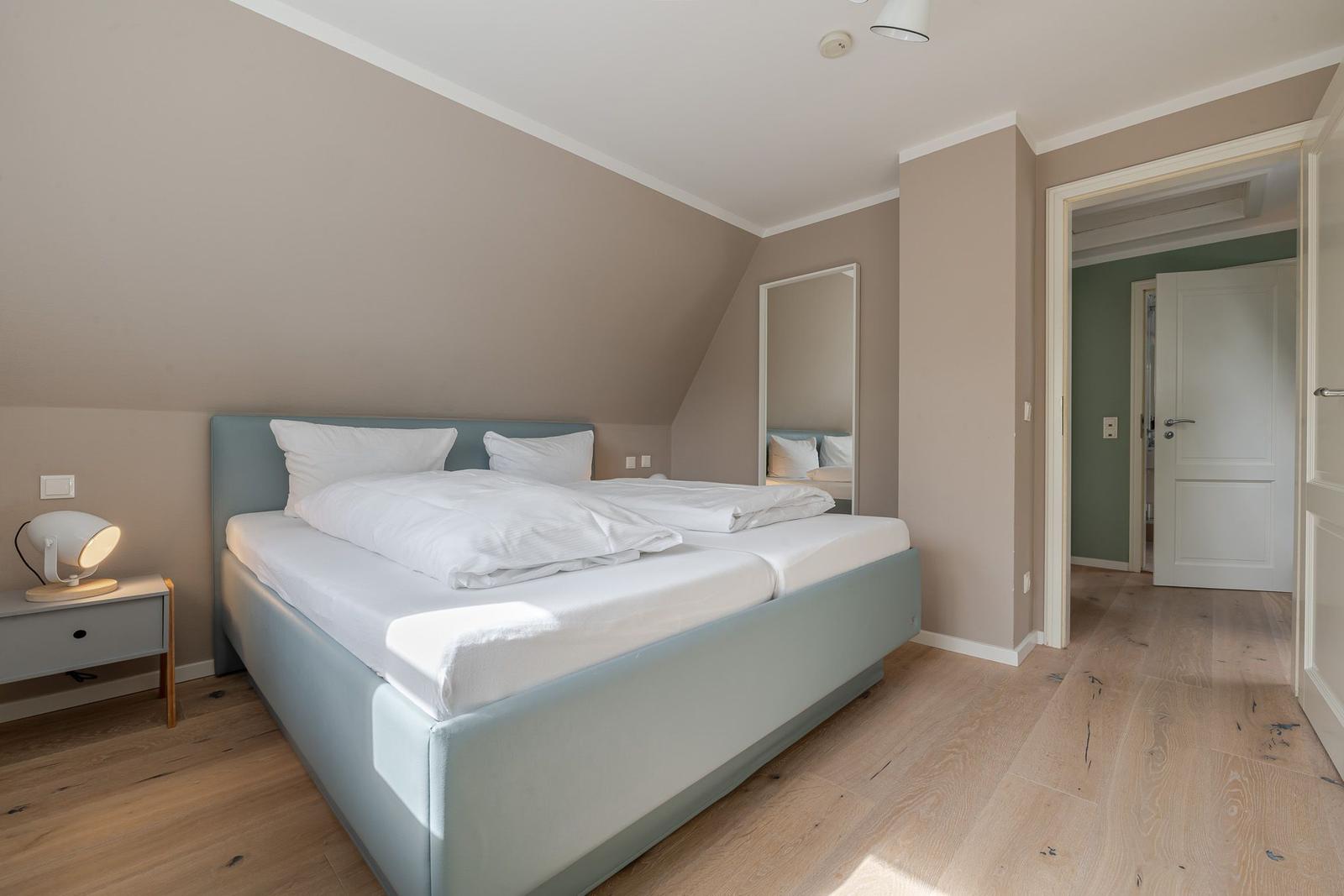 Schlafzimmer mit hellblauem Polsterbett, Nachttisch, Spiegel und Holzboden.