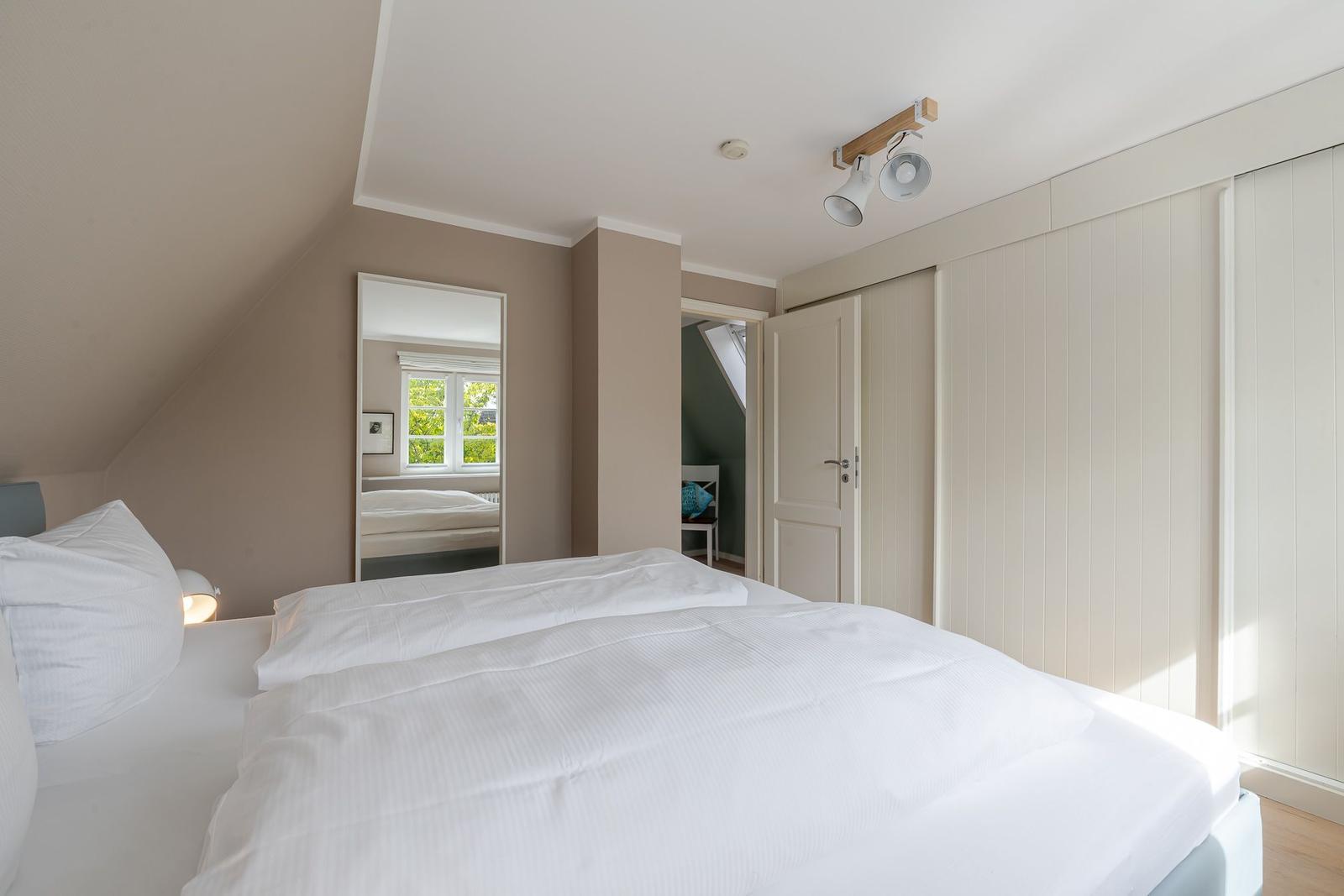 Schlafzimmer mit Dachschräge, weißem Doppelbett, Einbauschrank und Spiegel.