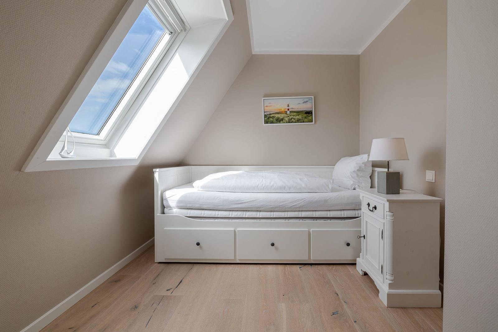 Dachschlafzimmer mit weißem Bett, Nachttisch, Lampe und Dachfenster.