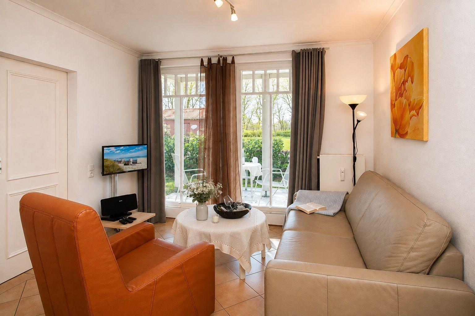 Wohnzimmer mit Sofa, orangefarbenem Sessel und TV. Durchgang zur Terrasse mit Blick auf Garten.