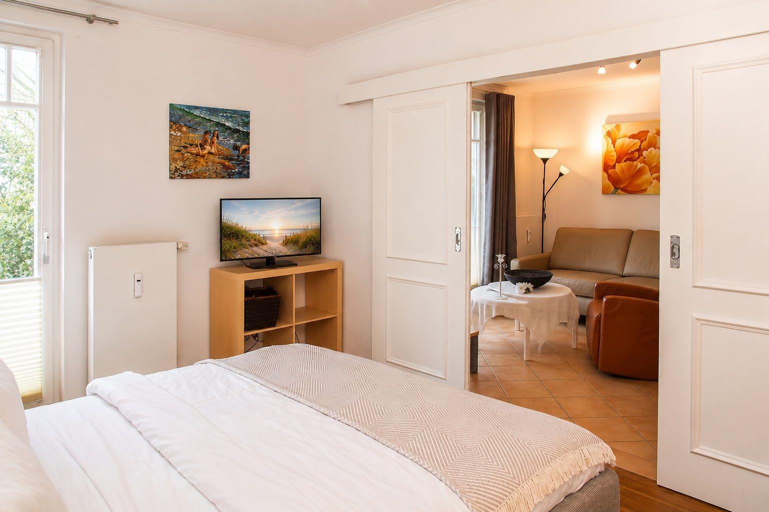 Schlafzimmer mit Bett und Fernseher. Durchgang in Wohnbereich mit Sofa und Beistelltisch.