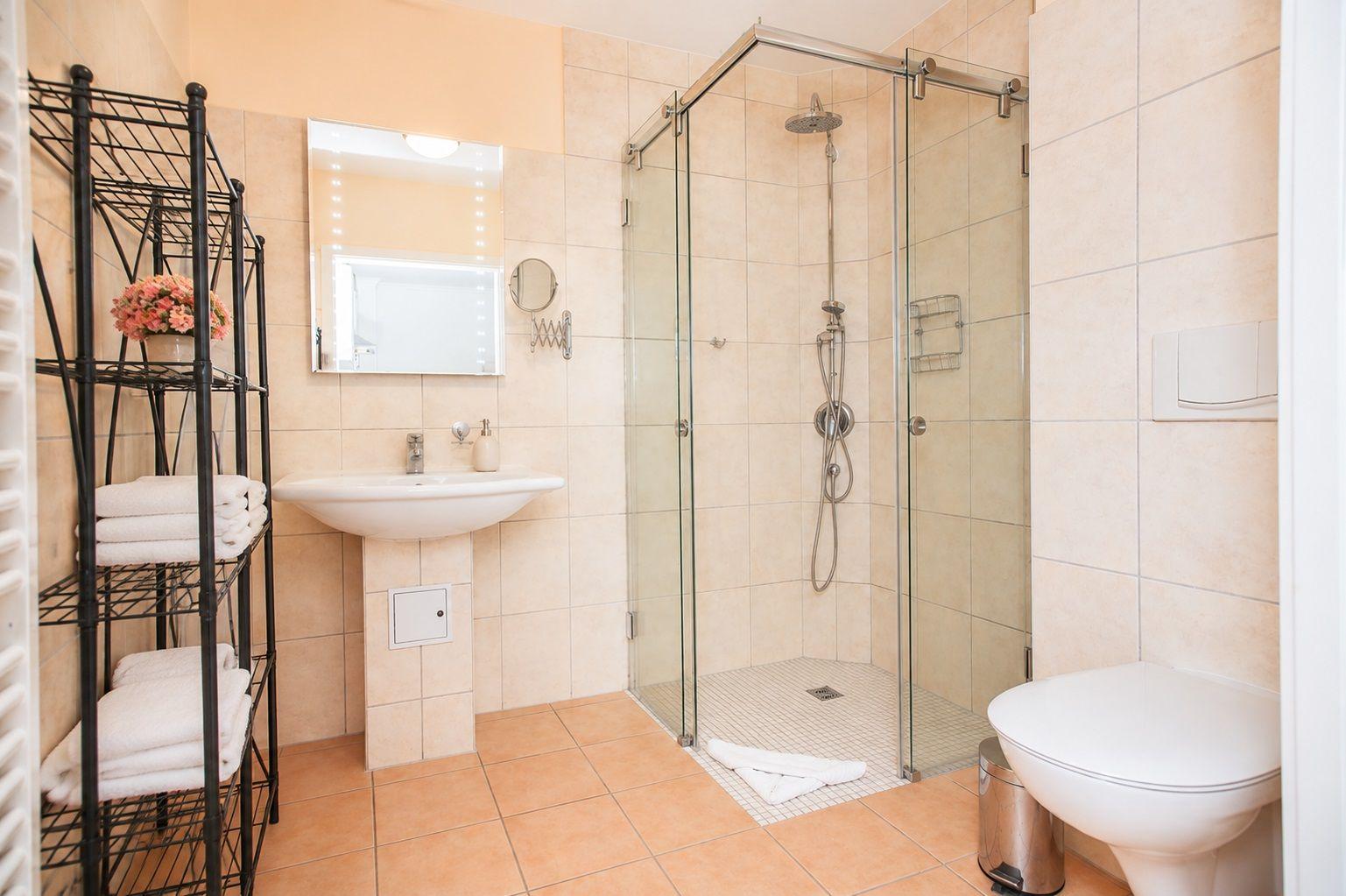 Badezimmer mit Dusche, Waschbecken und Toilette. Fliesenboden und Spiegel mit Beleuchtung.