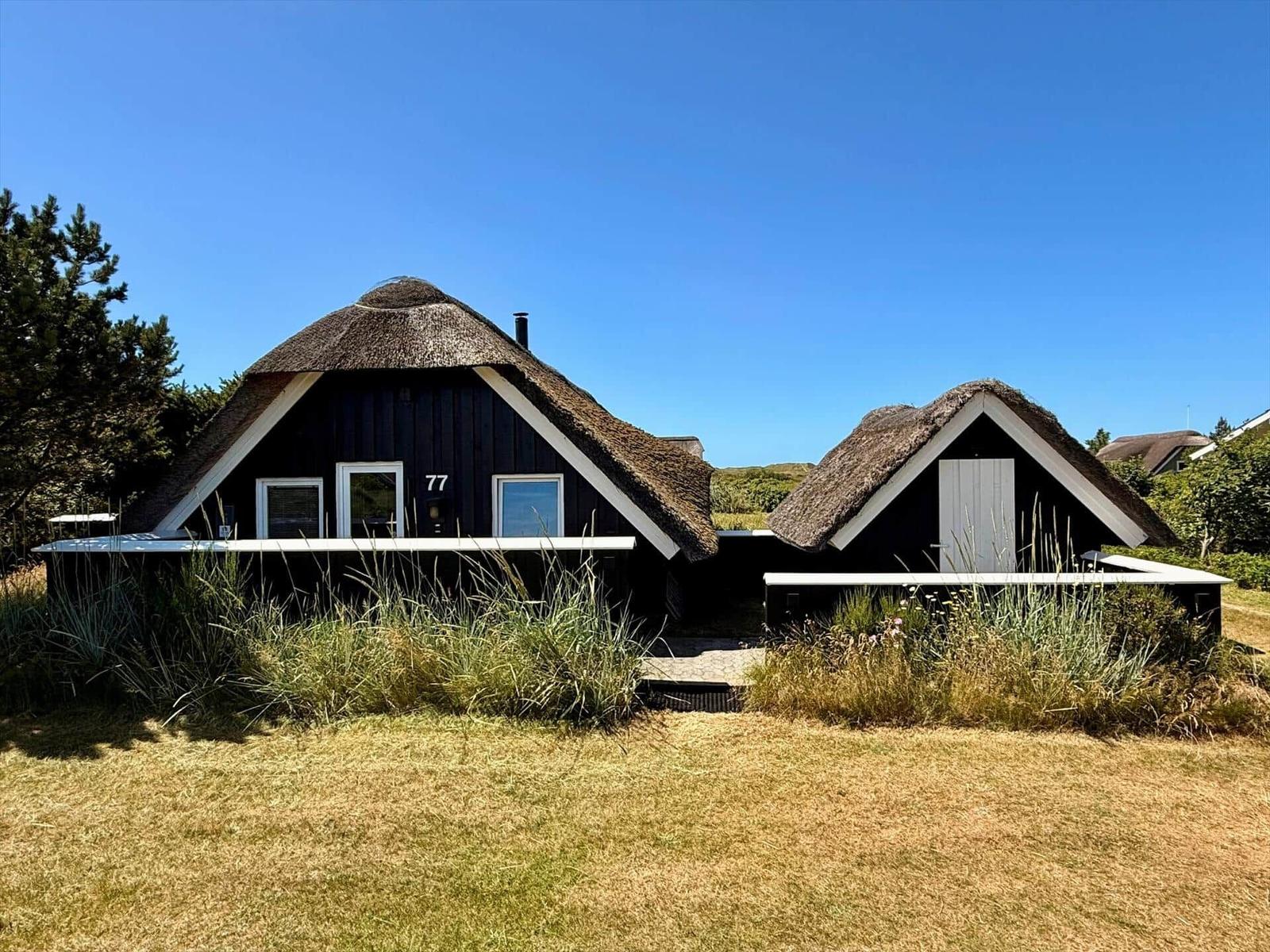 Haus mit Strohdach und Glasfront in grüner Umgebung unter blauem Himmel.