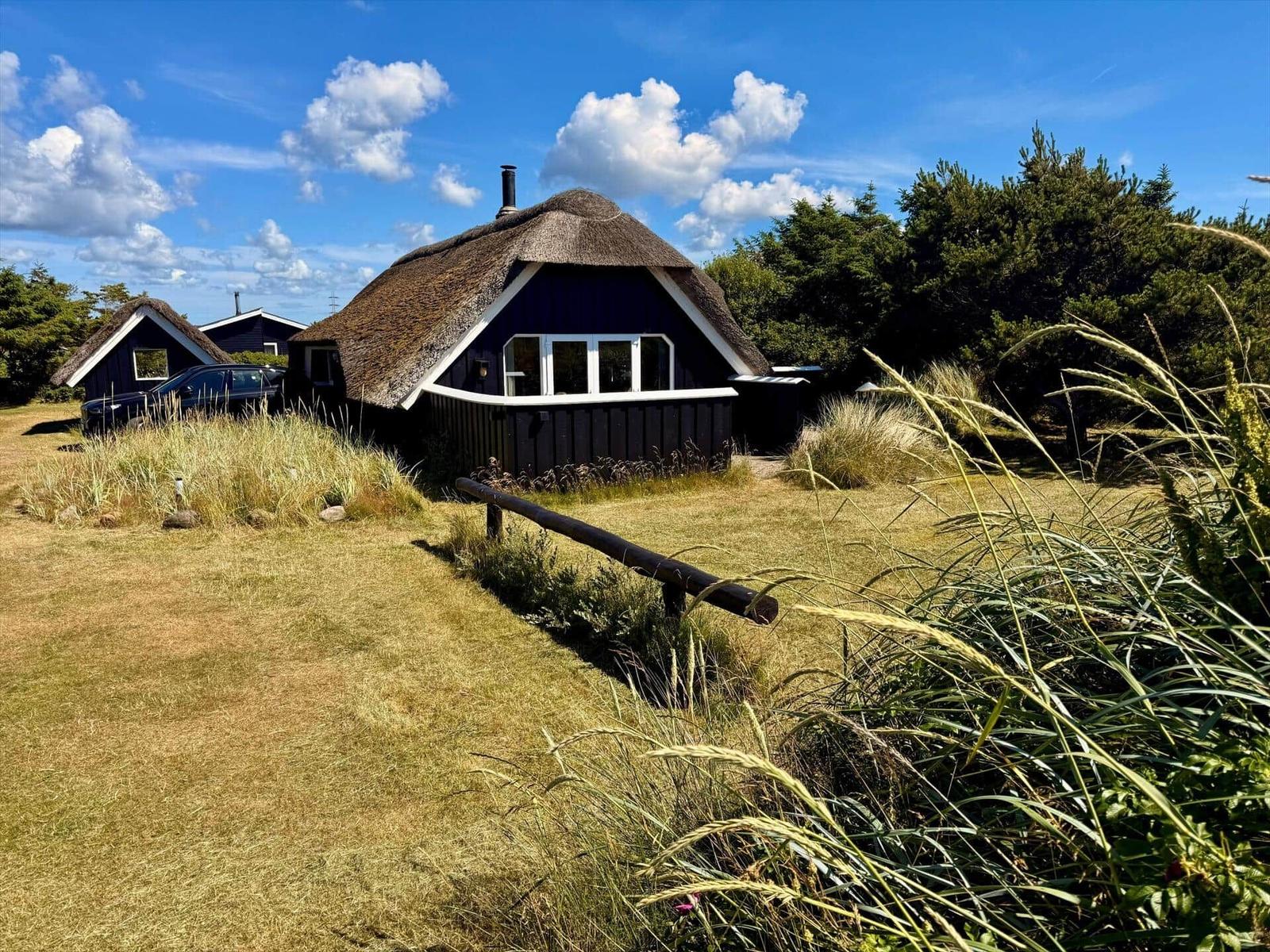 Dorfhaus mit strohgedecktem Dach und Fenstern, umgeben von Gras und einem Leuchtturm im Hintergrund.