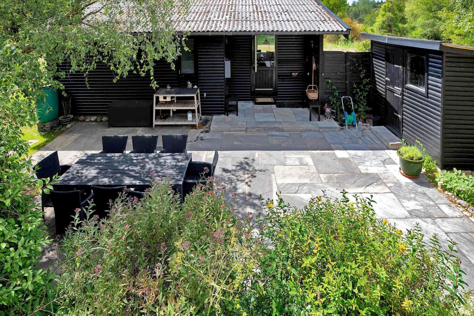 Schwarze Holzhütte mit Garten, Bank und Holzstapel. Steine führen zum Eingang.