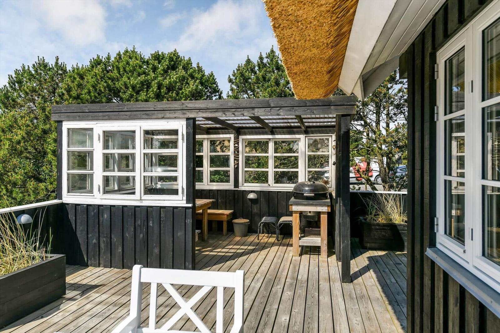 Holzterrasse mit weißer Bank vor einem Haus mit Strohdach und dunkler Fassade.