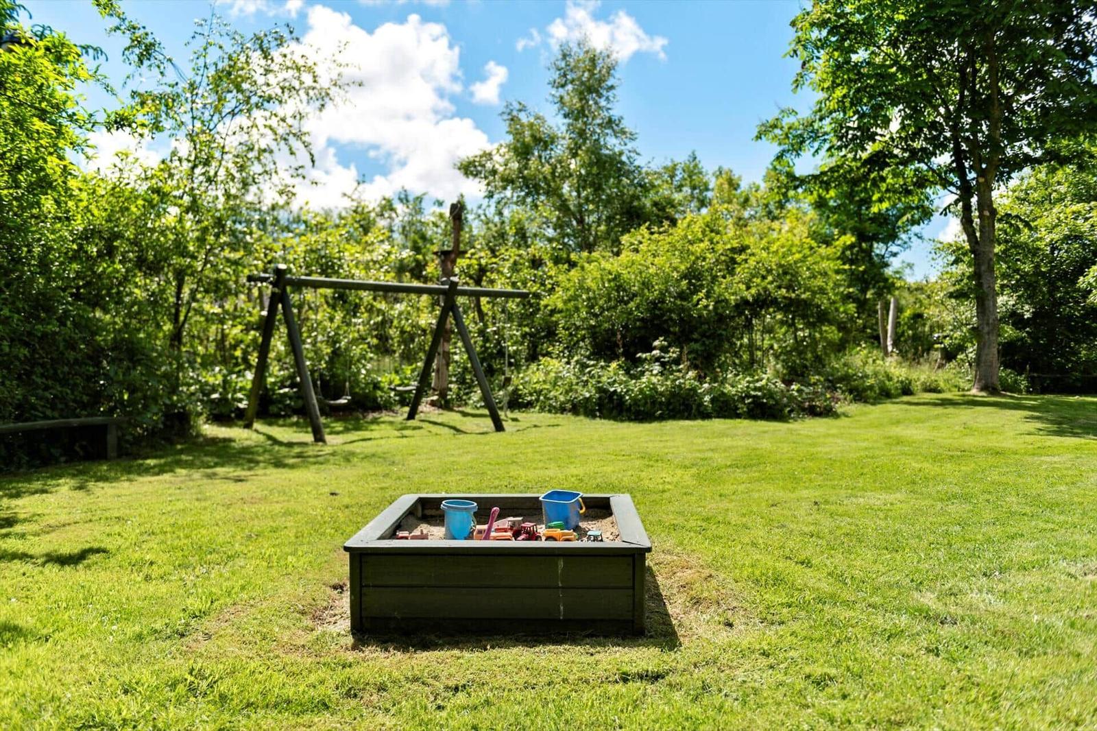 Rotes Holzhaus mit Garten und Sandkasten unter blauem Himmel.
