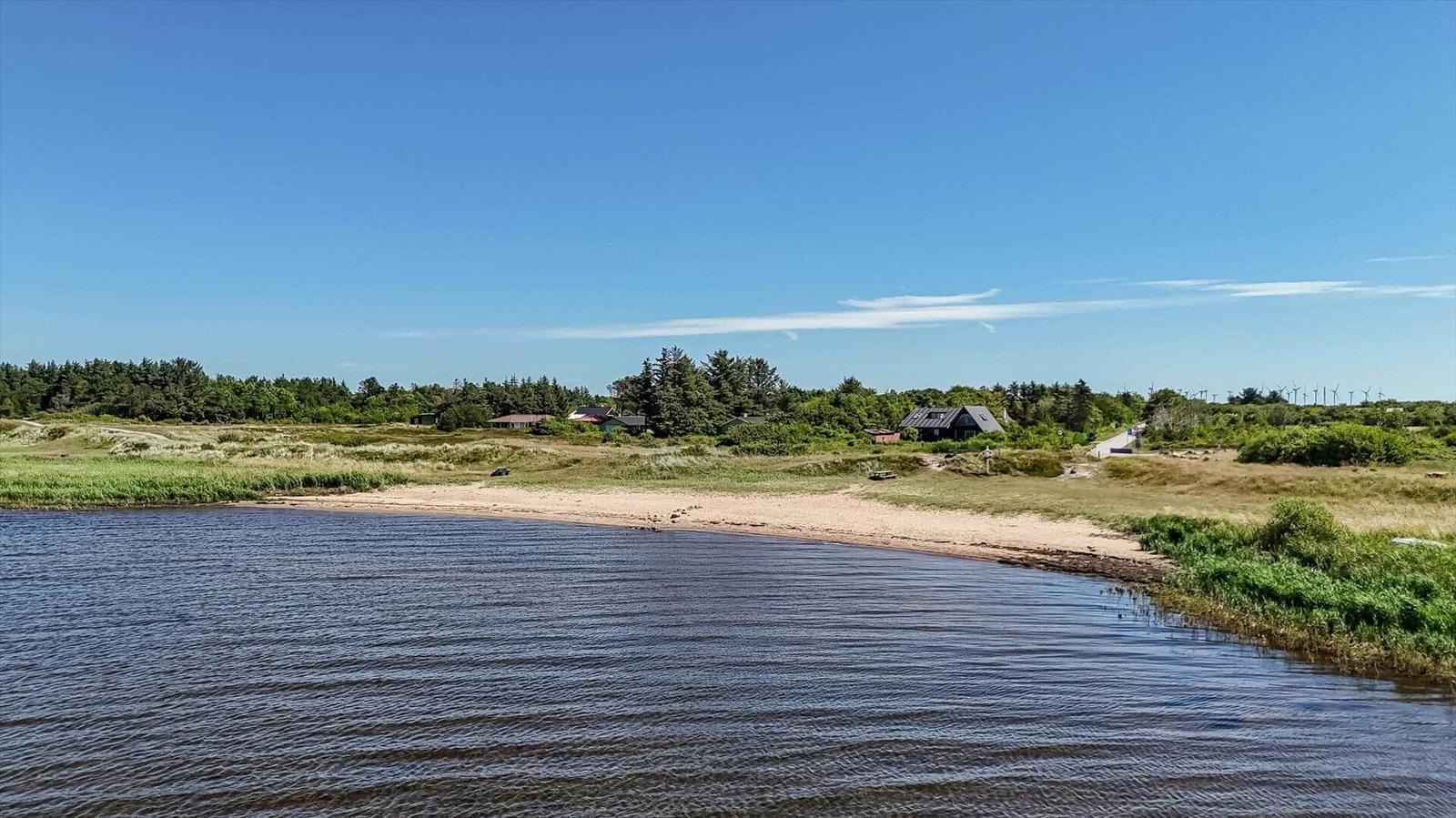 Strand mit Sand, Steinen und Gras am Wasser. Im Hintergrund sind Windräder sichtbar.
