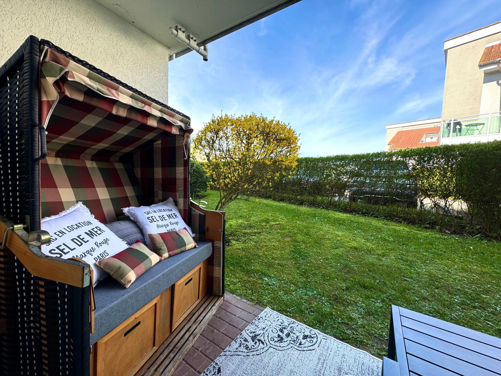 Terrasse mit Strandkorb, grünem Rasen und Blick auf Hecke und Gebäude.