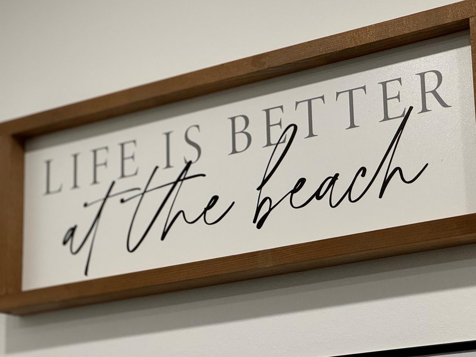 Ein Schild mit der Aufschrift Life is better at the beach in einem Holzrahmen an der Wand.