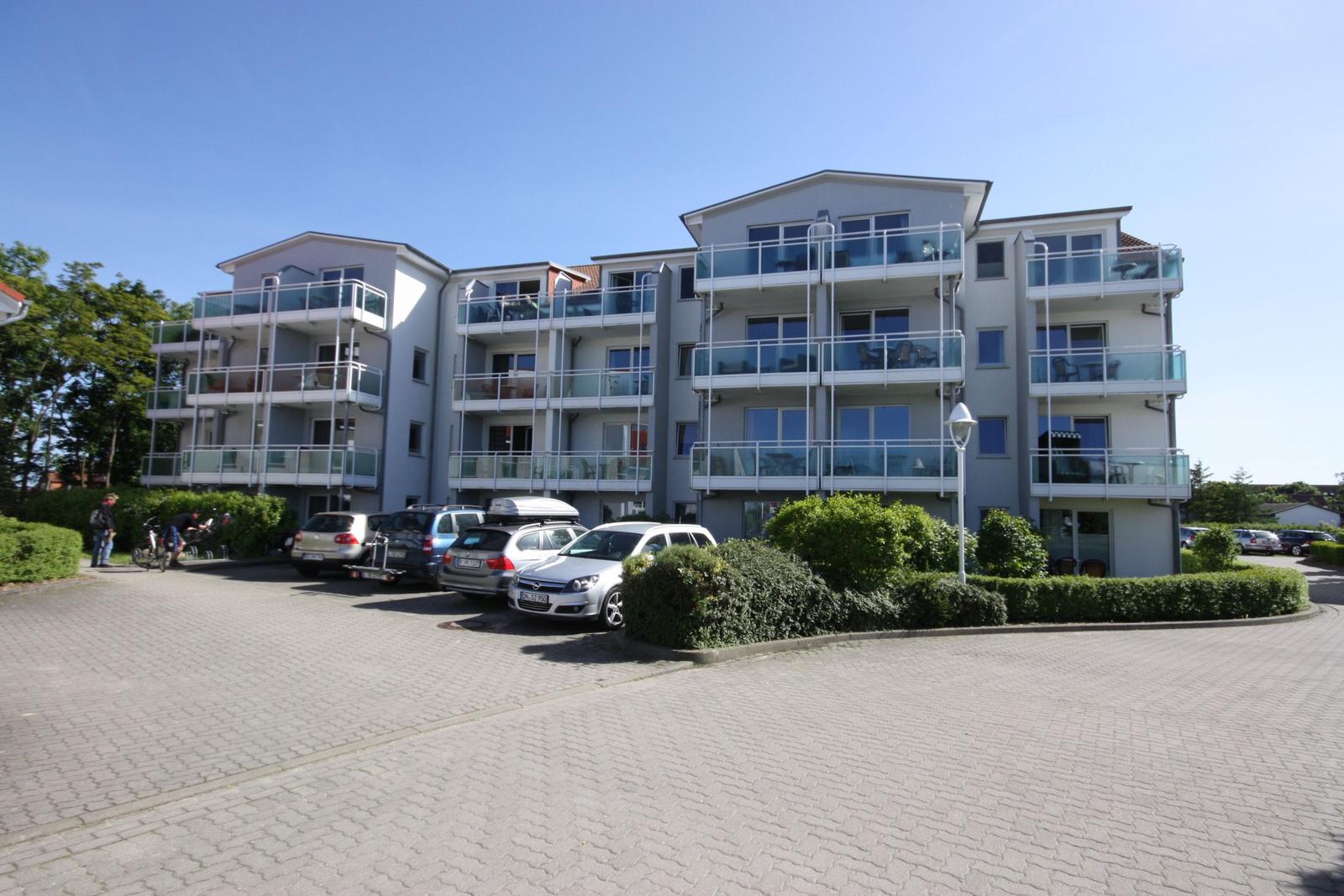 Wohnhaus mit Balkonen und gepflegtem Gartenbereich. Parkplatz und Ladesäule im Vordergrund.