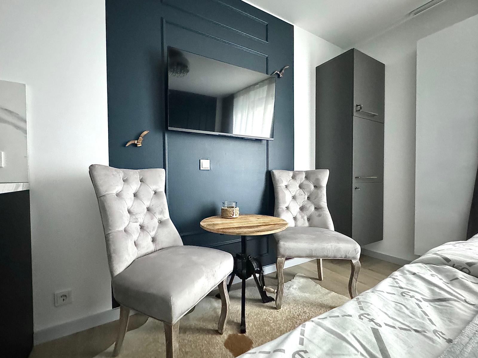 Zimmer mit TV, zwei Stühlen und kleinem Tisch vor dunkelblauer Wand.