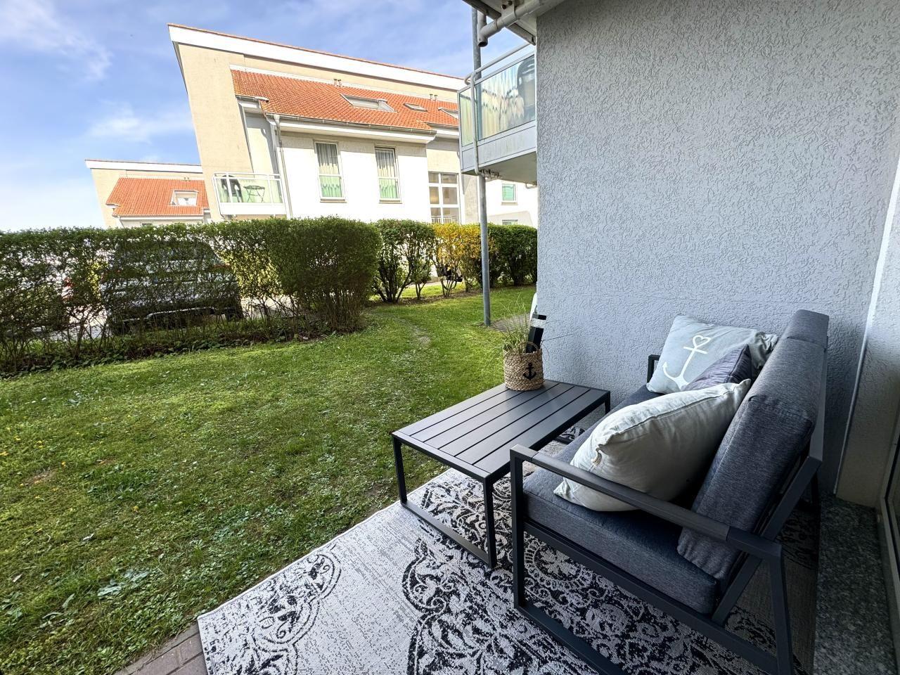 Terrasse mit Sitzgruppe, Tisch und Teppich. Grünflächen und Gebäude im Hintergrund.
