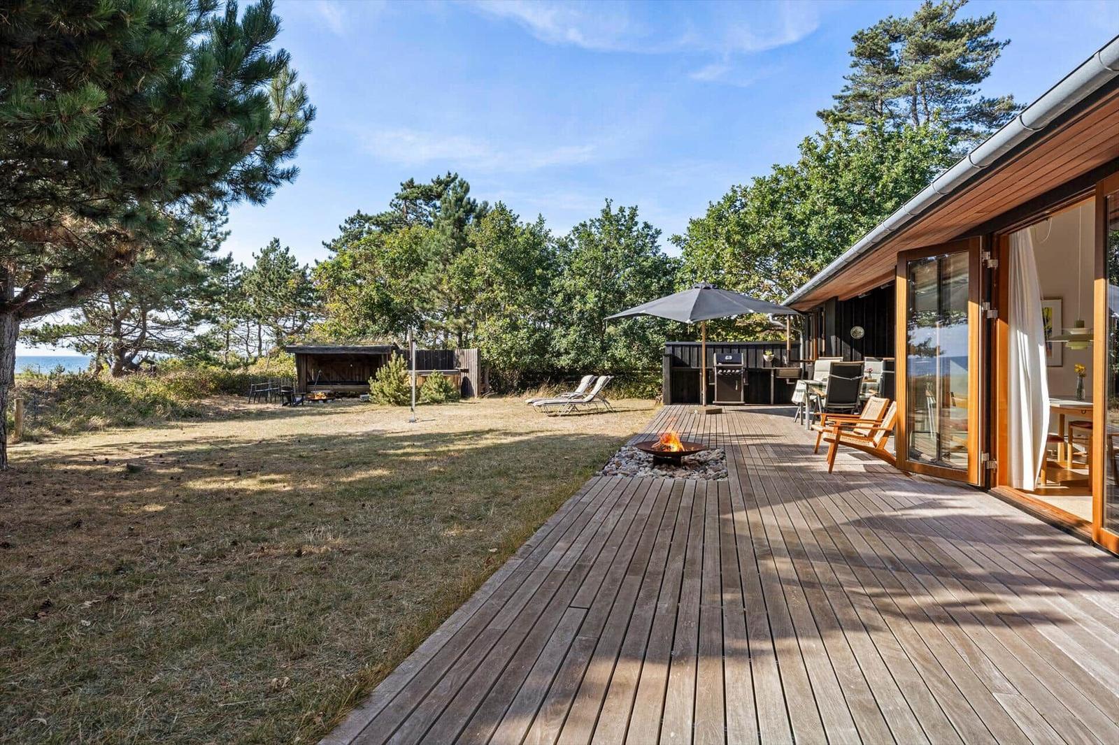 Wooden deck with fire pit, grill, armchairs and lawn, water in the background.