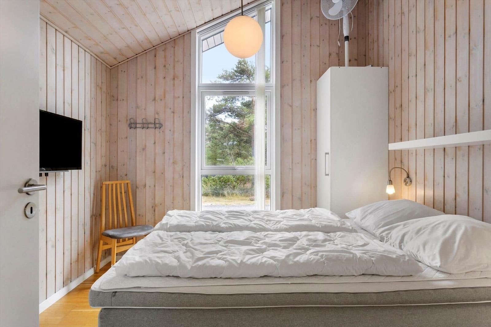 Schlafzimmer mit Bett, weißem Schrank, Holzstuhl, Wand-TV und schrägem Fenster.