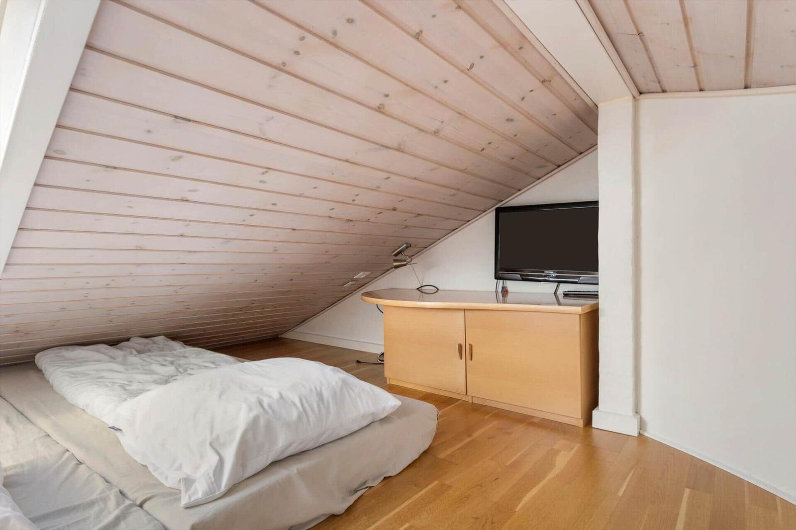 Schlafnische unterm Holzdach mit Matratze am Boden und Fernseher auf Kommode.