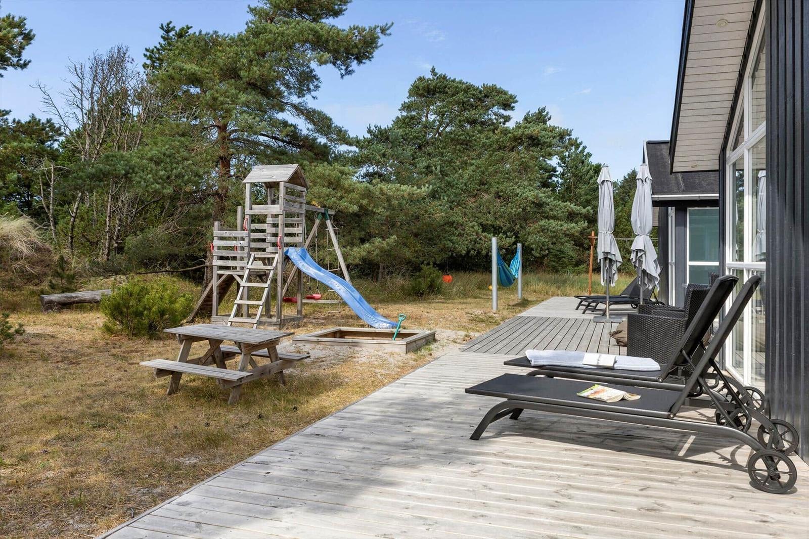 Holzterrasse mit Sonnenliegen, Picknicktisch, Hängematte und Spielgerät mit Rutsche.
