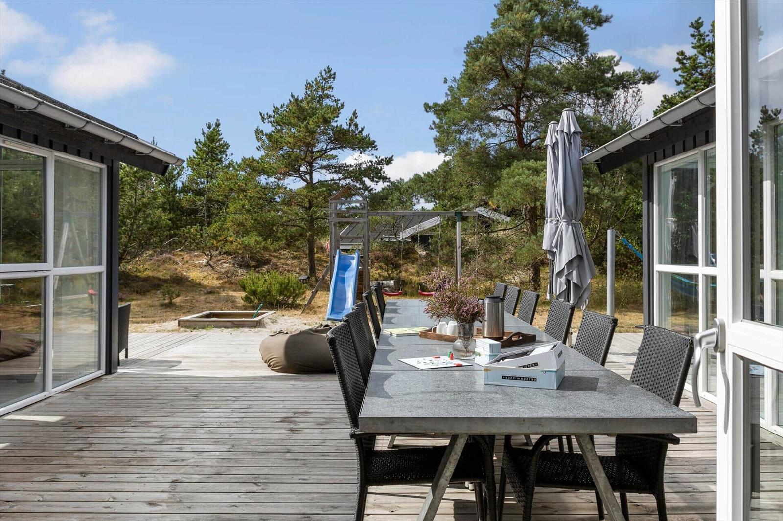 Holzterrasse mit Steintisch, schwarzen Stühlen und Sitzsack vor Spielgerät und Kiefern.