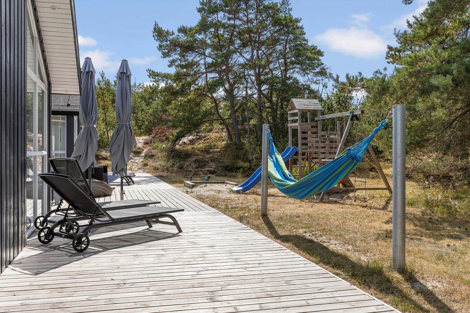 Holzterrasse mit Sonnenliegen, Schirmen, Hängematte und Holzspielgerät mit Rutsche.
