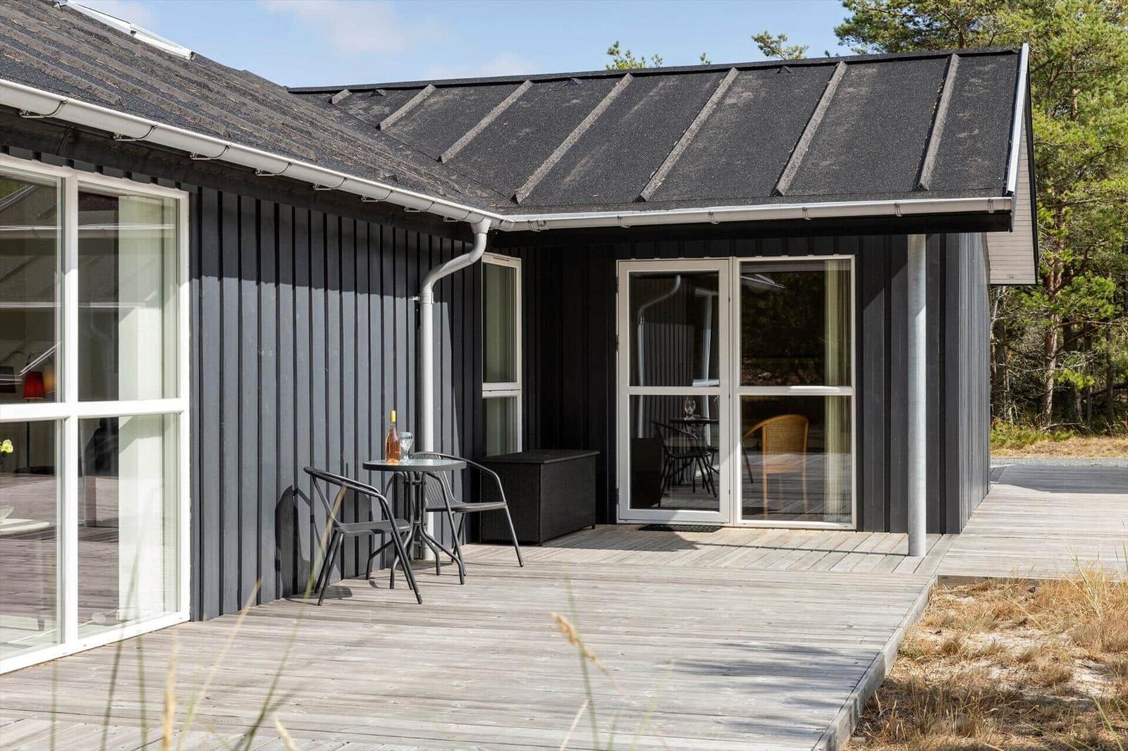 Holzterrasse am schwarzen Holzhaus mit rundem Tisch, zwei Stühlen und Glastür.