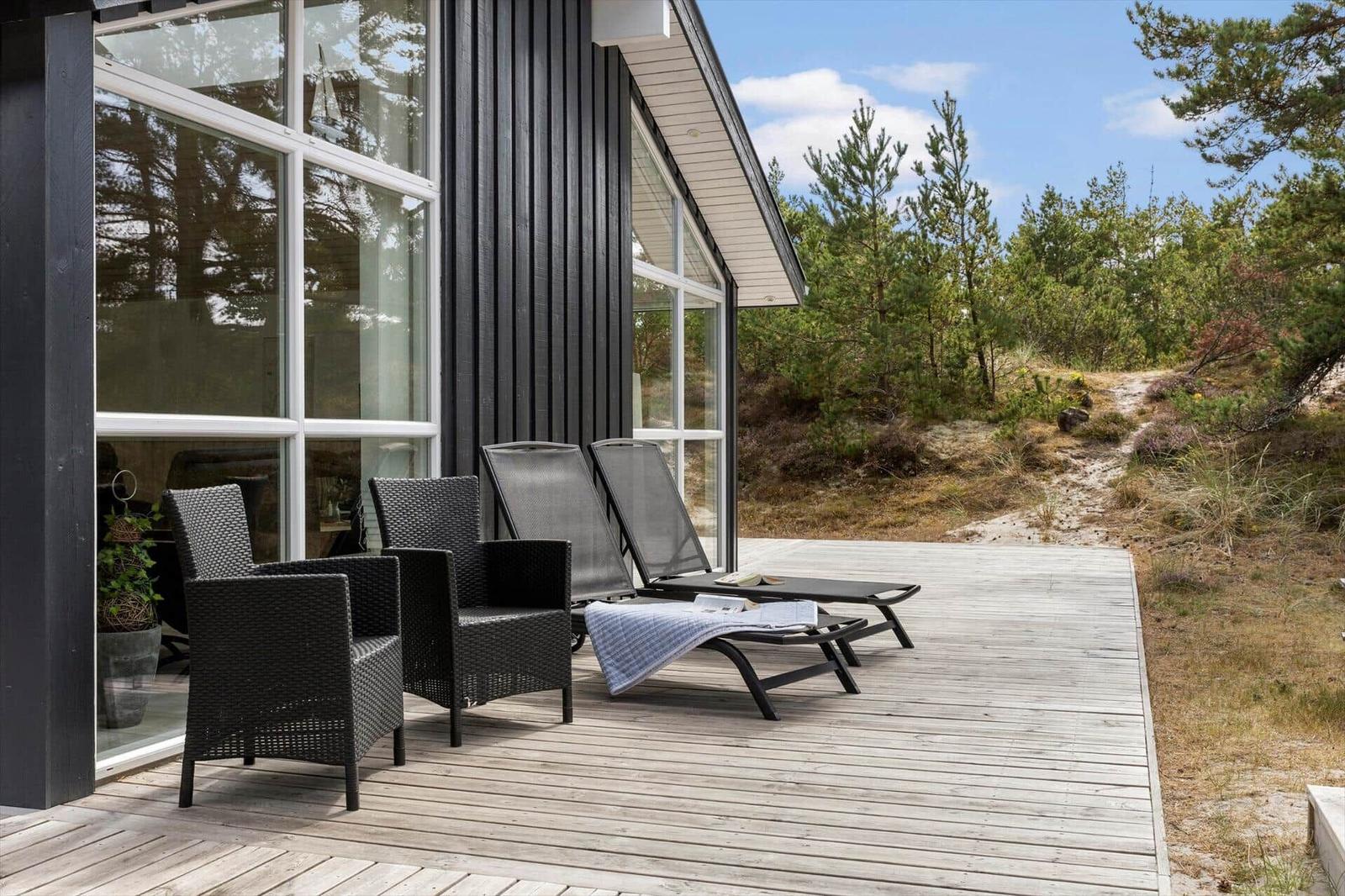 Holzterrasse mit Rattansesseln und Sonnenliegen am dunklen Haus vor Sand und Kiefern.