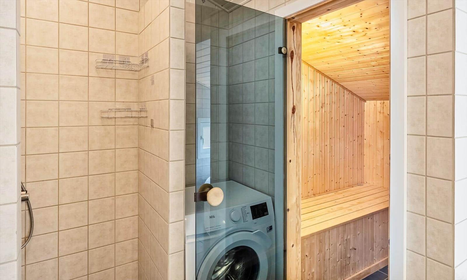 Geflieste Dusche mit Drahtablagen, Waschmaschine und offener Holzsauna.