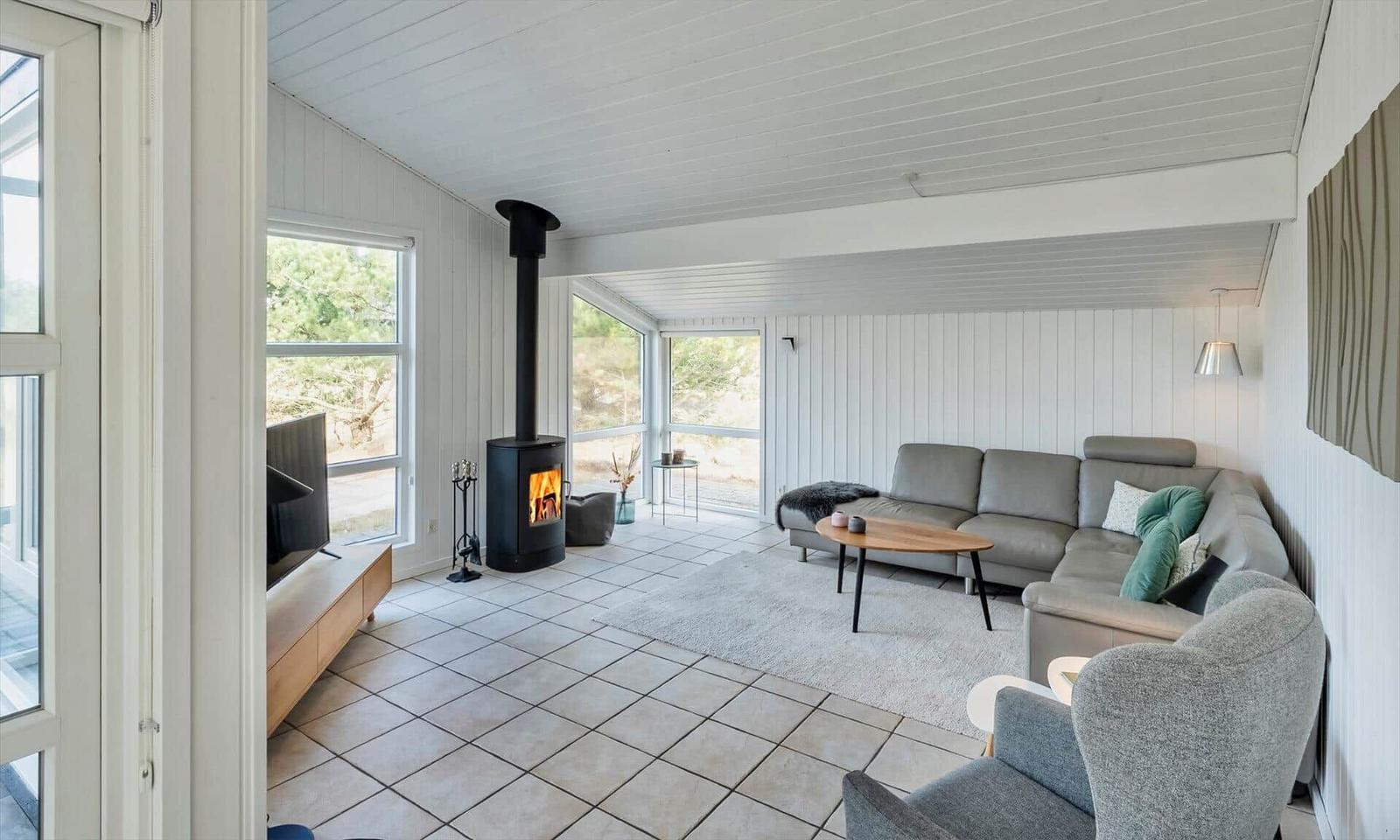 Wohnzimmer mit grauem Ecksofa, Kaminofen, Fernseher und großen Fensterfronten.