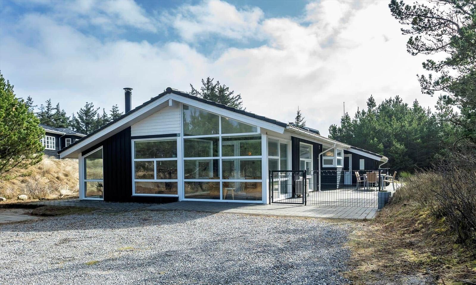 Schwarzes Holzhaus mit weißer Glasgiebelwand, Steinterrasse und Kiefern am Kiesweg.