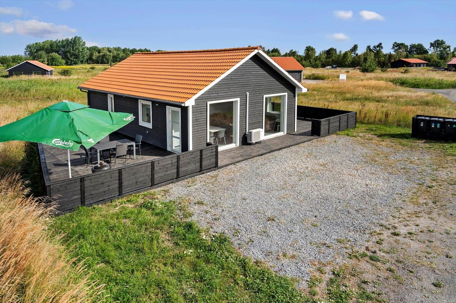 Dark wooden house with tiled roof, terrace, seating under a green parasol and gravel area.