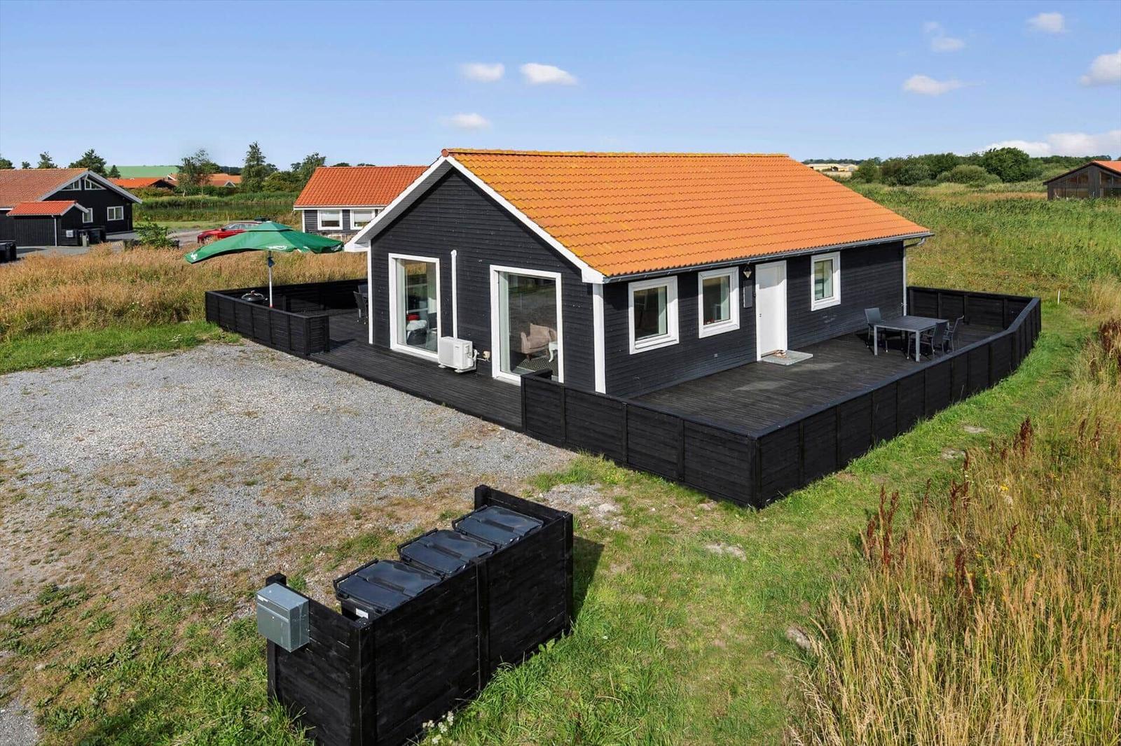 Black wooden house with large terrace, parasol and waste bins by a gravel area.