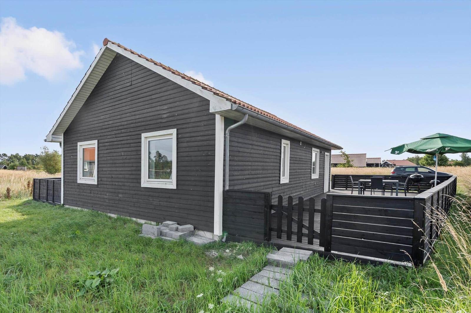 Dark wooden house with terrace gate, barbecue, parasol and paving slabs in the grass.