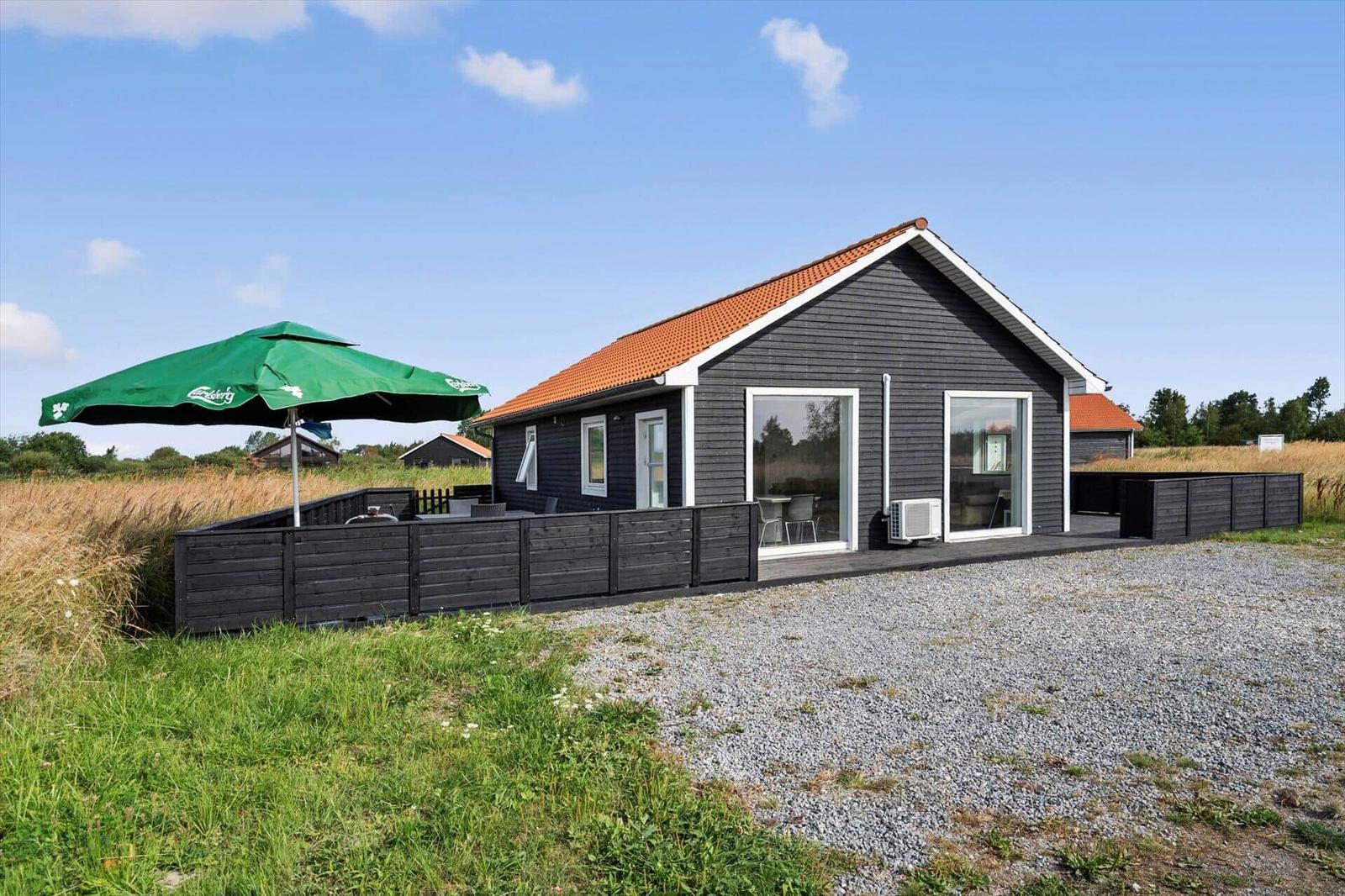 Black wooden house with large windows, outdoor AC unit, gravel area and green parasol.