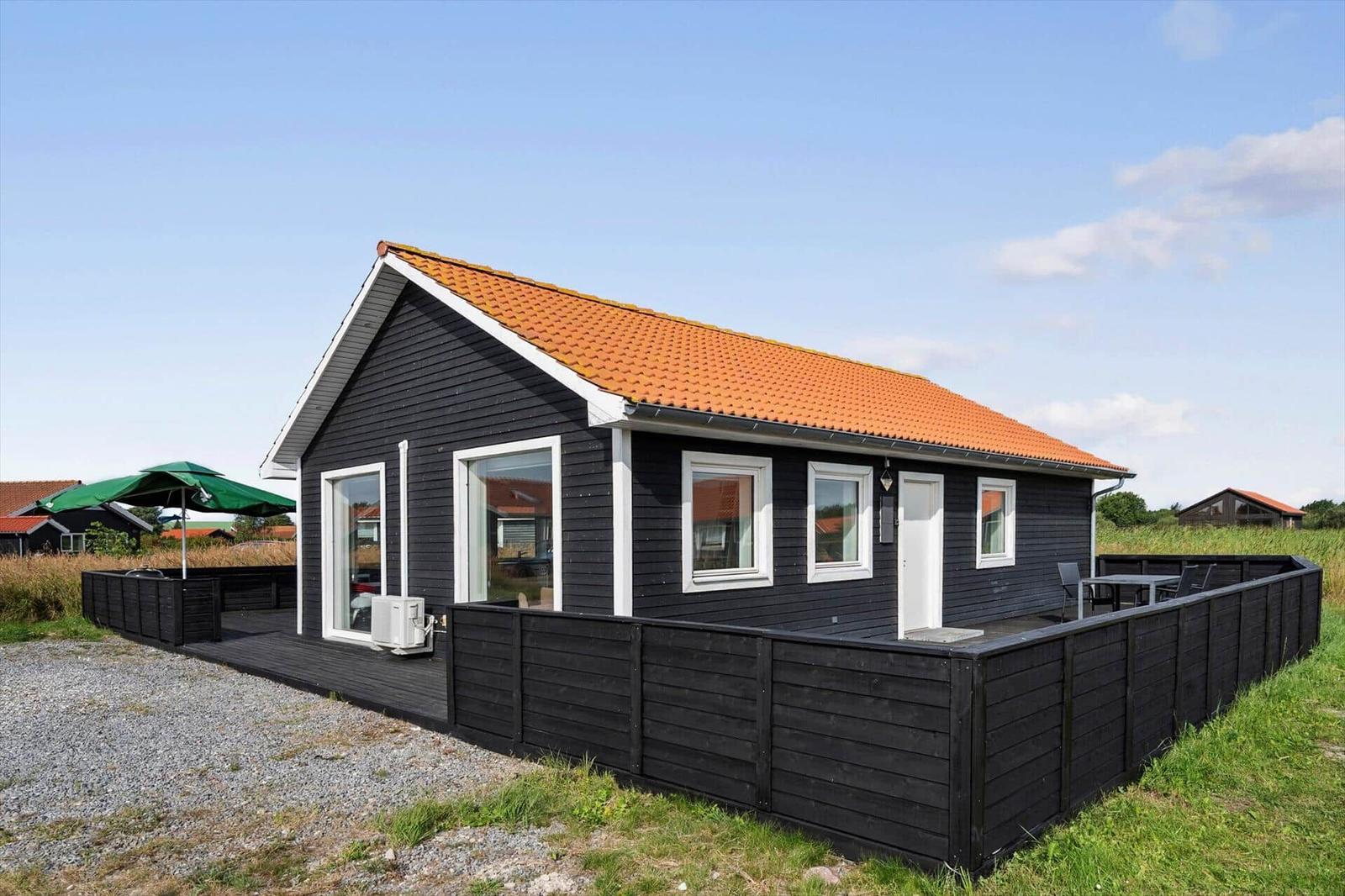 Black wooden house with red tiled roof, white door and terrace with a green parasol.