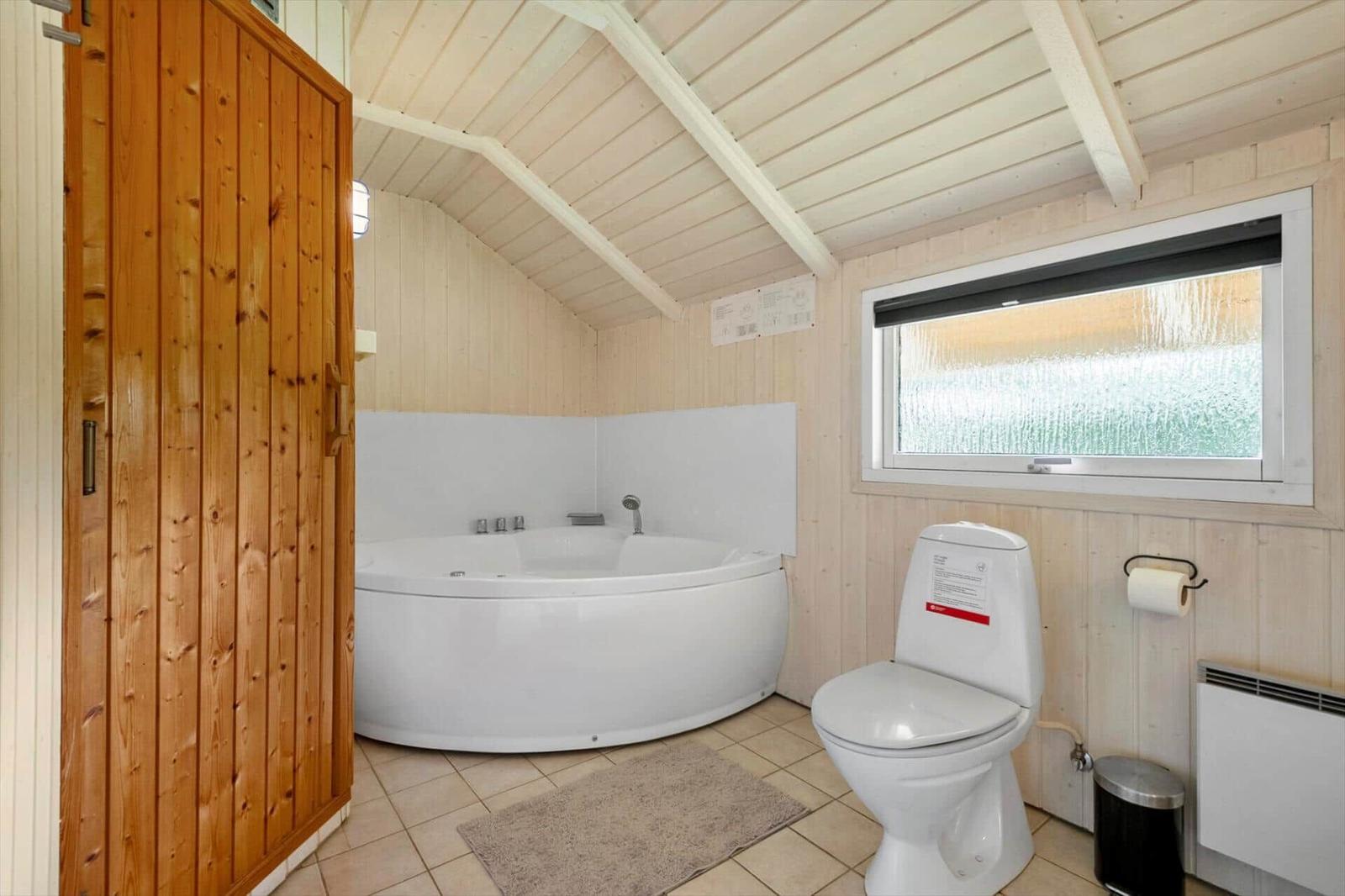 Bathroom with white whirlpool tub, toilet and sauna door under sloped wood ceiling.