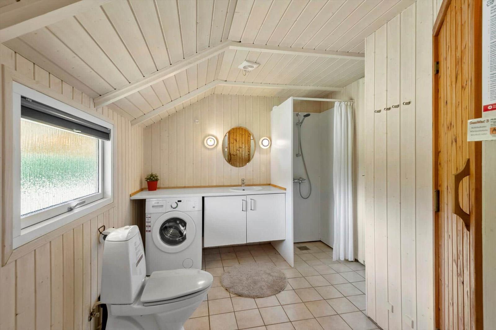 Bathroom with toilet, washing machine, round mirror, shower and wooden sauna door.