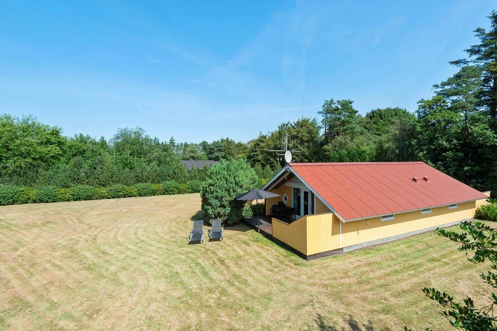 Yellow wooden house with red roof, terrace, parasol and sun loungers on the lawn.