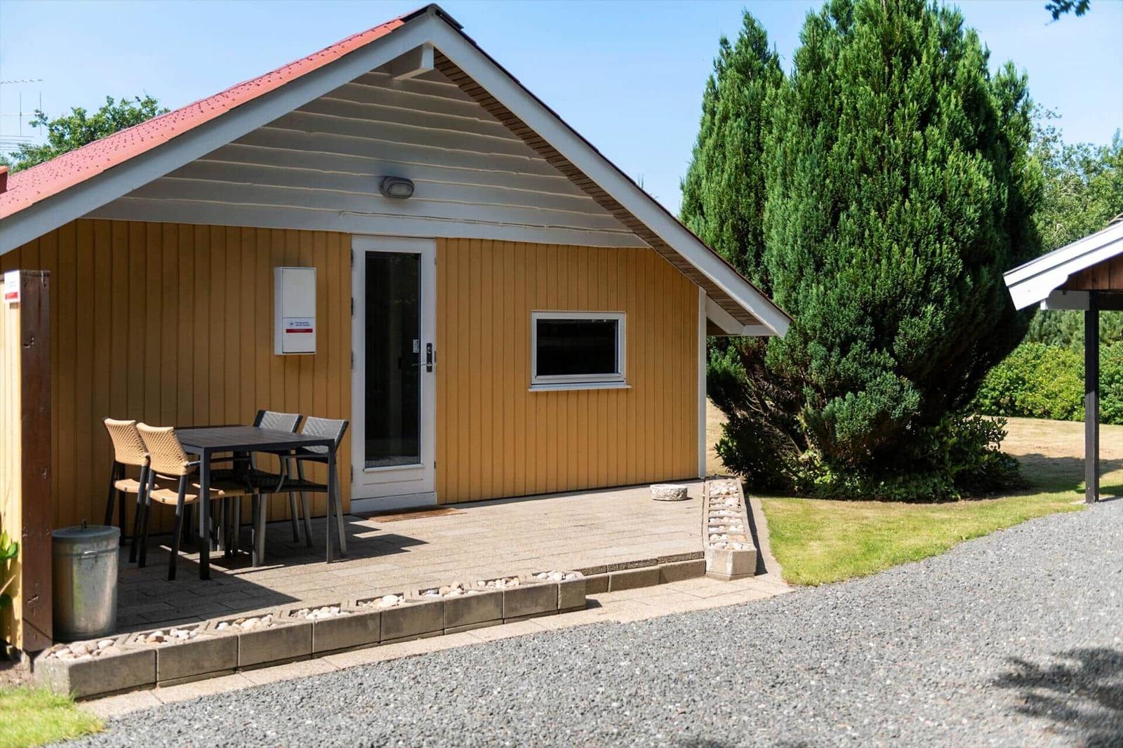 Yellow wooden house with glass door, stone patio, dining table and four garden chairs.