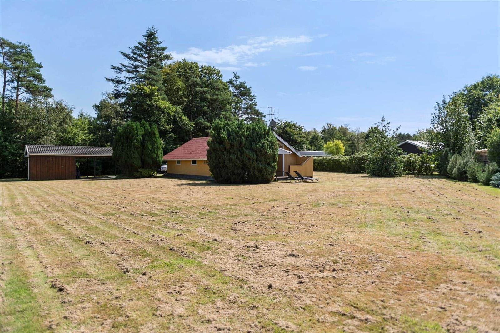 Yellow wooden house with red roof on a wide lawn beside a dark outbuilding.