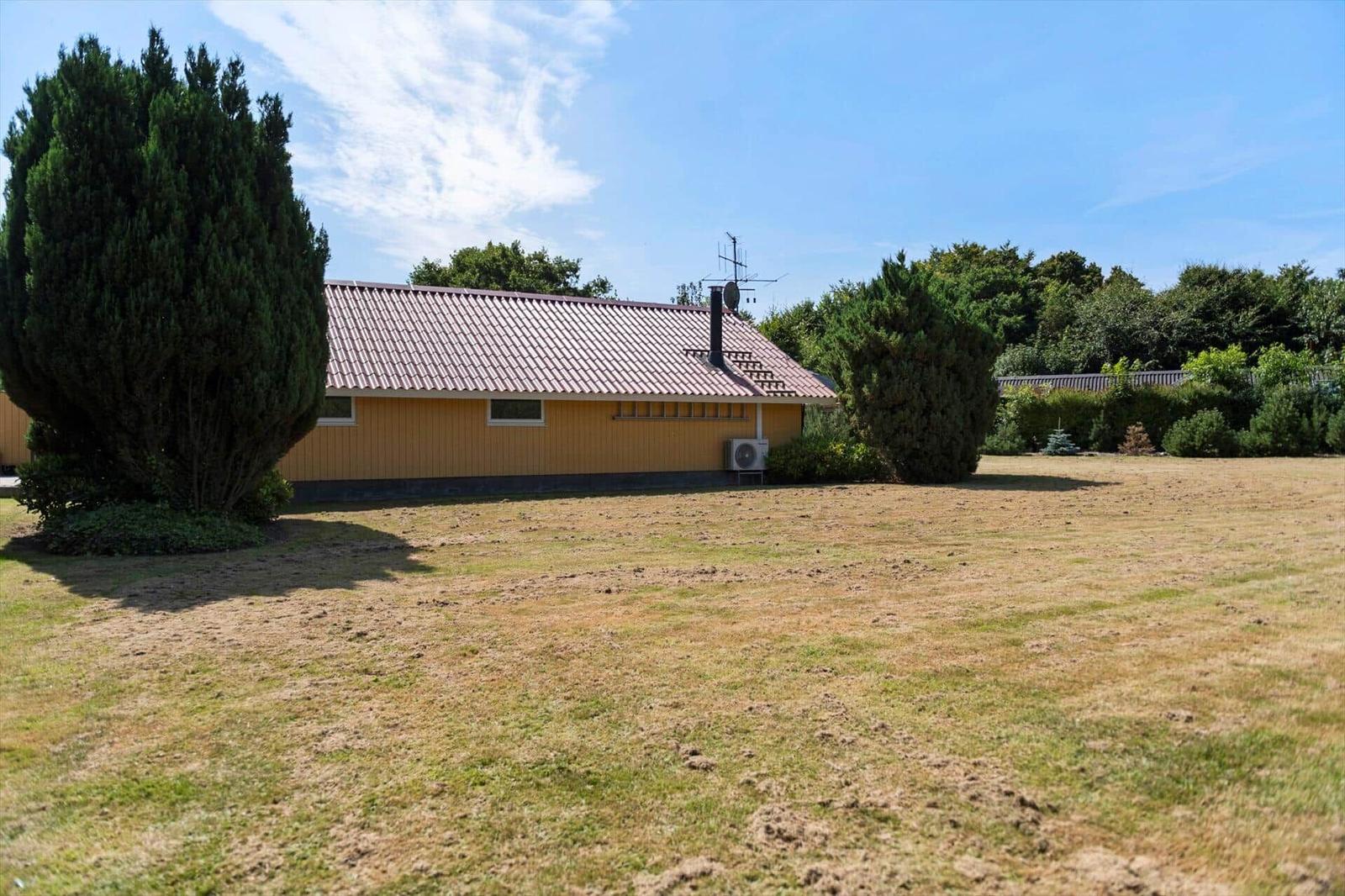 Yellow wooden house with red tiled roof on a lawn between tall trees.