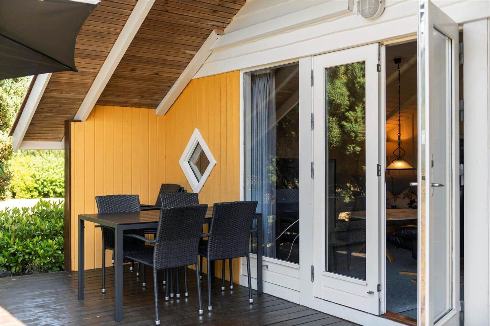 Covered terrace with dark dining table and chairs against a yellow wooden facade.