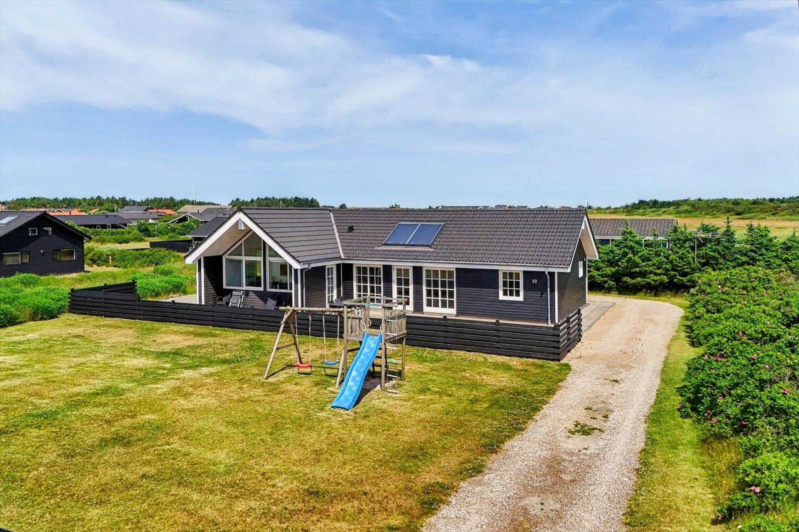 Luftbild eines Ferienorts mit Häusern, Grünflächen und Blick aufs Meer mit Windkraftanlagen.