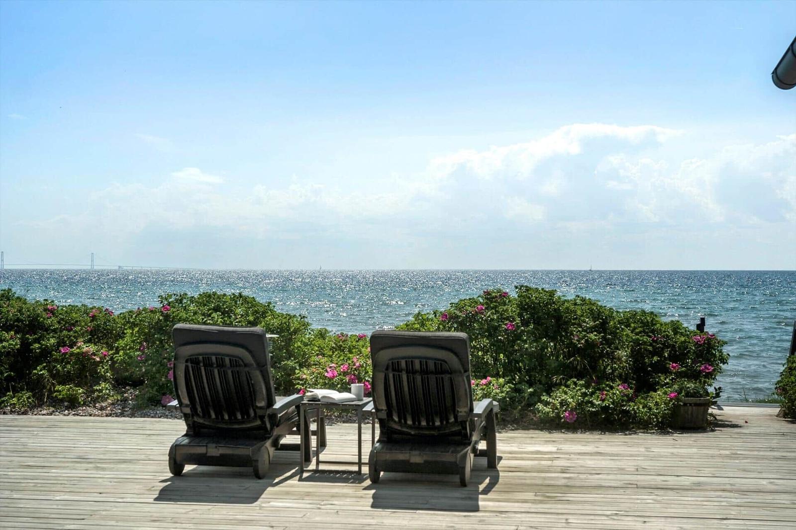 Terrasse mit Sitzgruppe und Tisch, Blick auf Meer und grüne Hecke mit rosa Blumen.