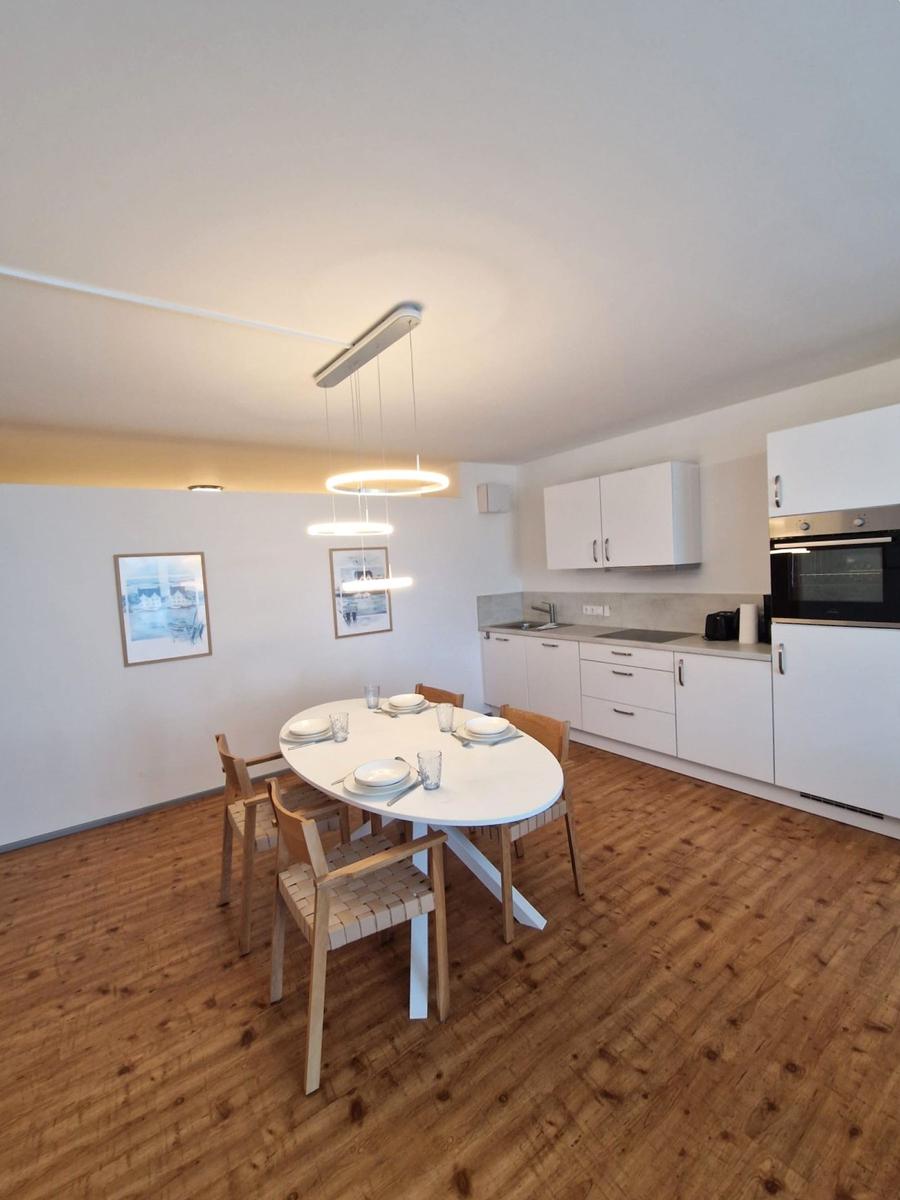 Ovaler Esstisch mit Holzstühlen und Ringlampe vor weißer Küchenzeile.