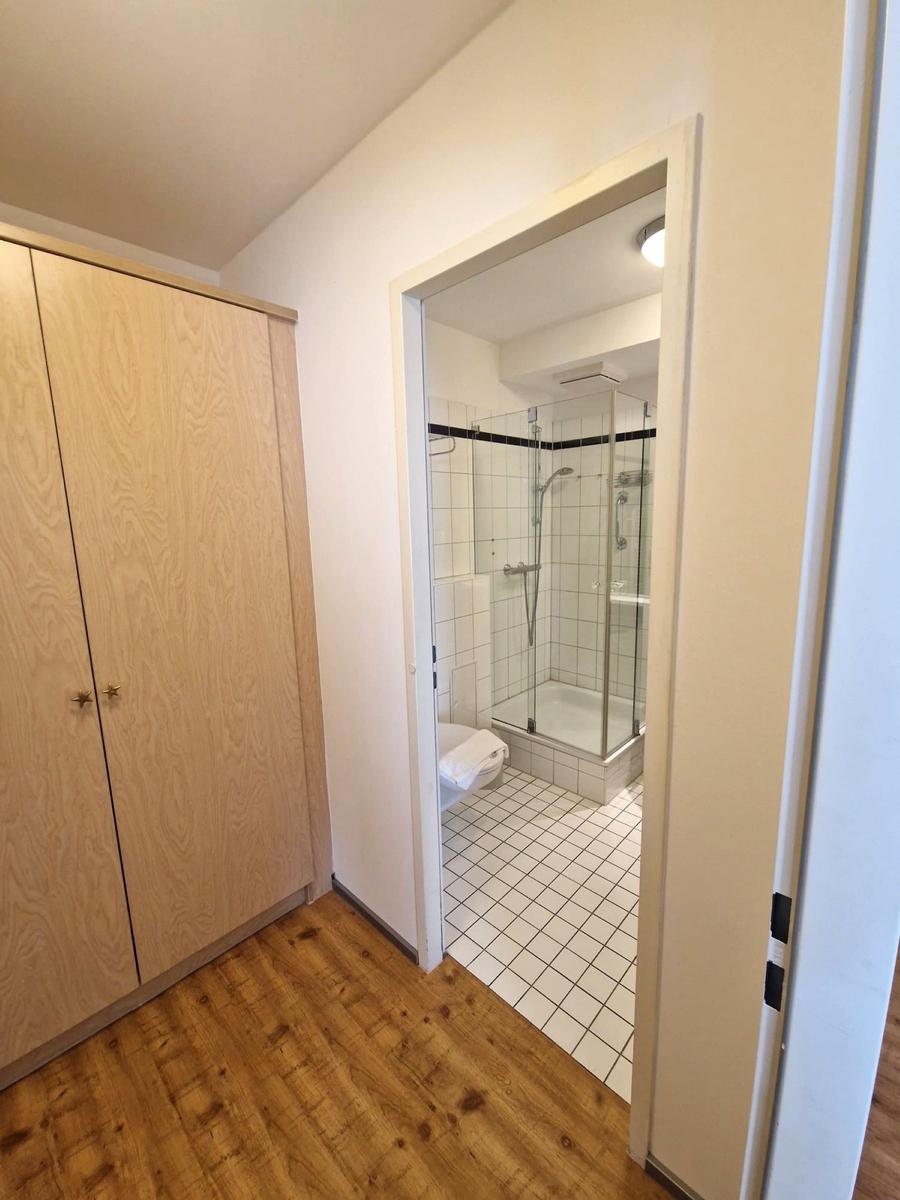Heller Holzschrank und Blick ins Bad mit Glasdusche und WC.