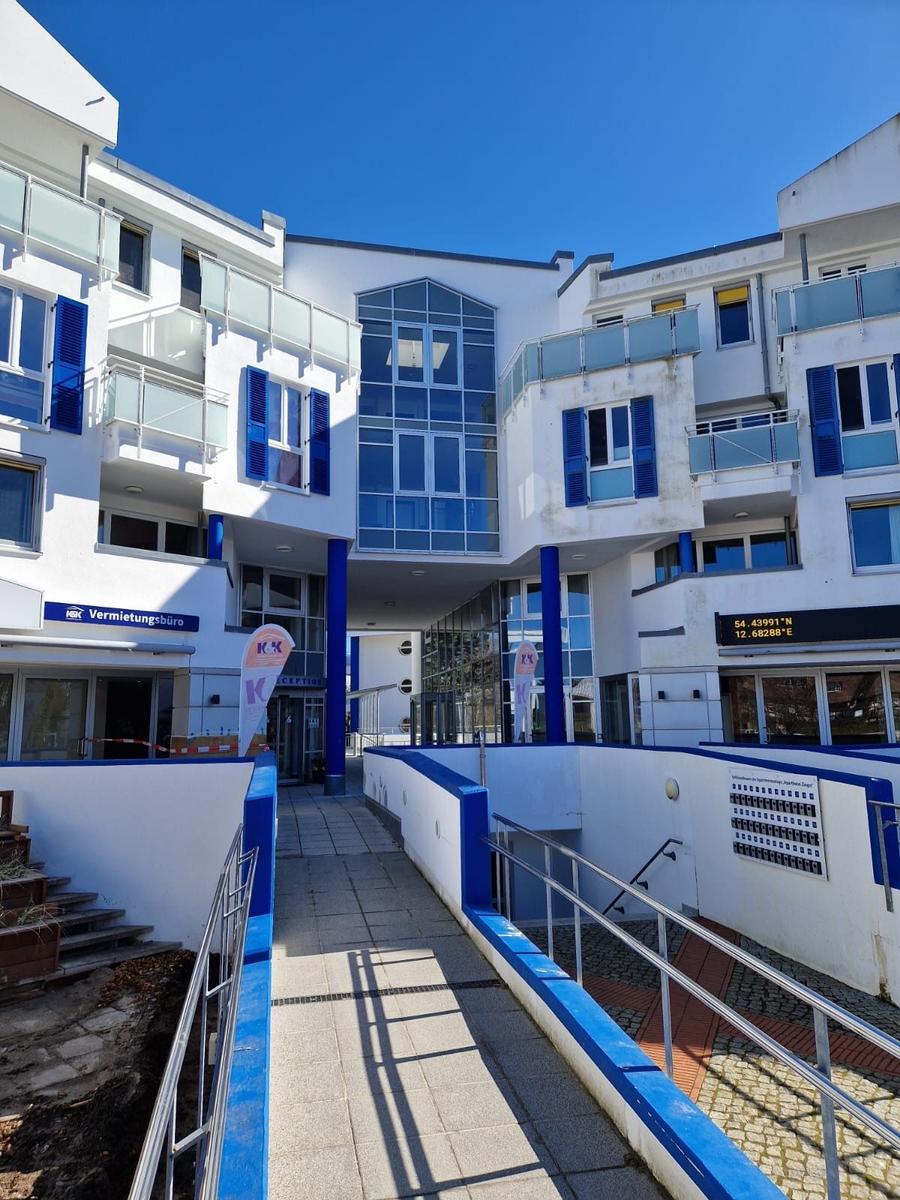 Weißer Wohnbau mit blauen Stützen, Glasbalkon und gepflastertem Hofzugang.