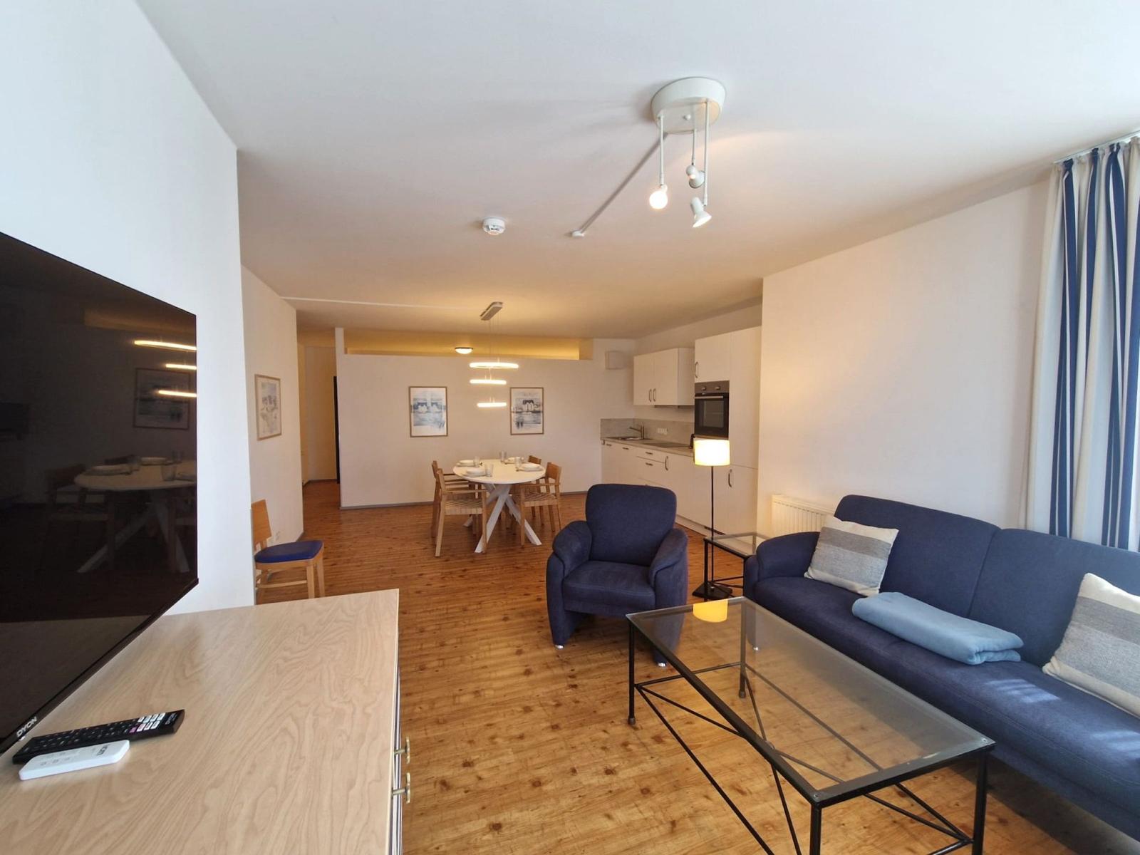 Wohnzimmer mit blauem Sofa, Sessel, Glastisch und Essbereich zur Küche.